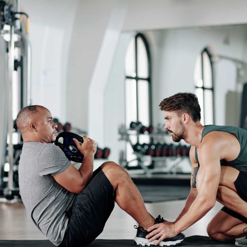 Young serious fitness trainer helping mature client doing sit-ups crunches on mat in gym